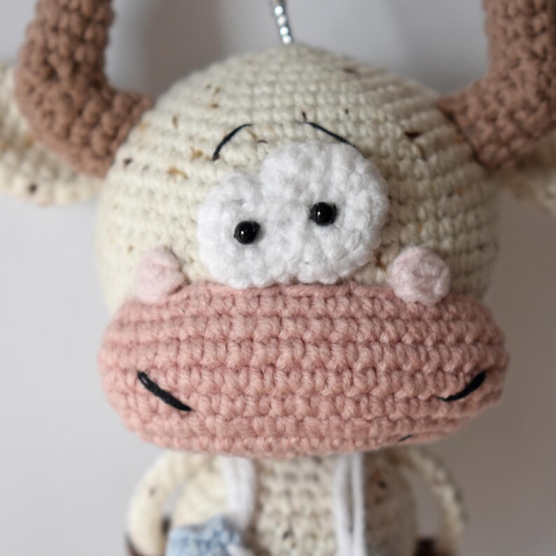 May include: A crocheted stuffed animal toy shaped like a cow with brown horns, white fur, and a pink nose. The cow has black eyes and a white face.