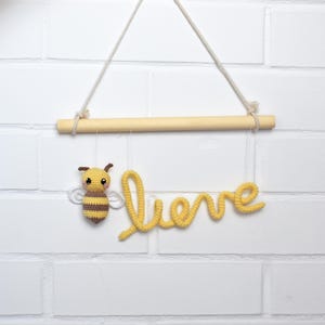 May include: A handmade crochet wall hanging featuring a yellow and brown bee and the word "believe" in yellow script. The sign hangs from a wooden dowel suspended by a natural rope against a white brick wall.