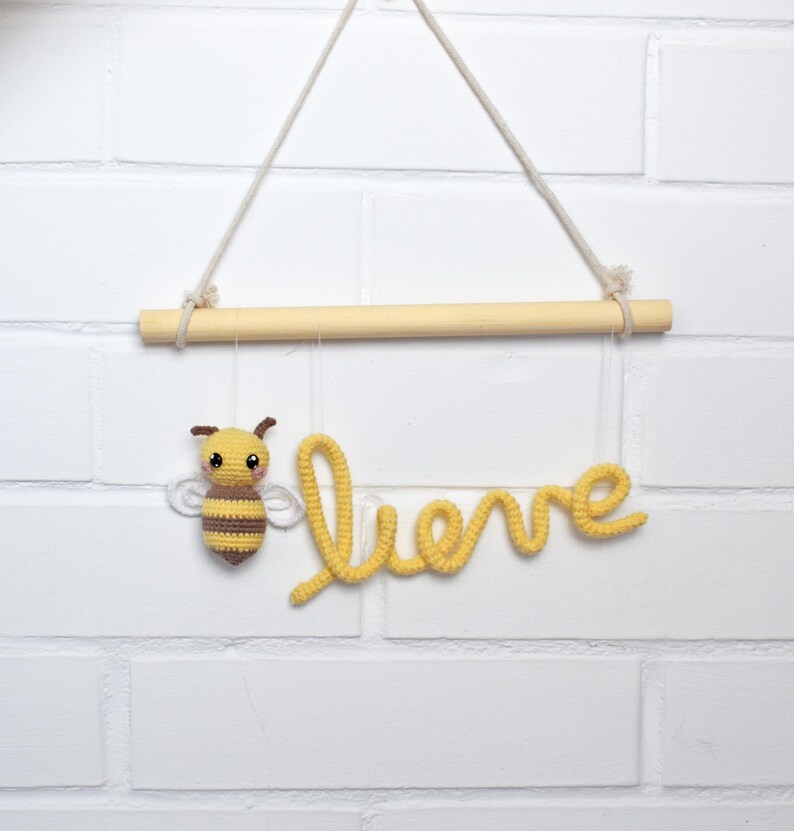 May include: A handmade crochet wall hanging featuring a yellow and brown bee and the word "believe" in yellow script. The sign hangs from a wooden dowel suspended by a natural rope against a white brick wall.