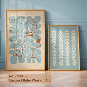 Peut inclure: Deux impressions d'art encadrées. L'une présente le profil d'une femme dans des tons bleu clair et orange, avec un motif floral. L'autre impression a un fond bleu clair avec des motifs de feuilles blanches. Les deux sont dans des cadres dorés.