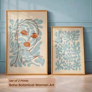 Peut inclure: Deux impressions encadrées. L'une représente un visage de femme avec des éléments botaniques bleus et bruns. L'autre présente un motif botanique bleu sur fond crème. Les cadres sont dorés. Le texte en bas indique "Set of 2 Prints Boho Botanical Woman Art."