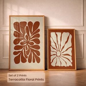 Peut inclure: Deux impressions florales en terracotta encadrées. La plus grande présente un motif floral stylisé dans des tons bruns sur fond crème. La plus petite a un motif similaire en crème sur fond marron. Les cadres sont de couleur marron clair. Le texte en bas indique "Set of 2 Prints" et "Terracotta Floral Prints."