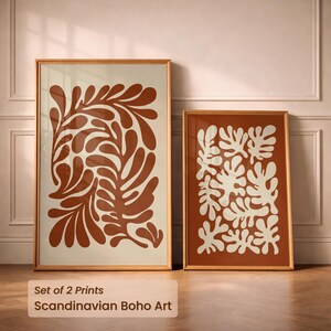Peut inclure: Deux impressions d'art encadrées avec des motifs botaniques abstraits. L'une présente un motif couleur rouille sur fond crème, l'autre un motif crème sur fond rouille. Les cadres sont dorés. Le texte en bas indique "Set of 2 Prints Scandinavian Boho Art."