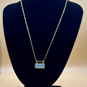 May include: A gold chain necklace with a light blue rectangular pendant. The pendant has a gold frame and a textured surface.