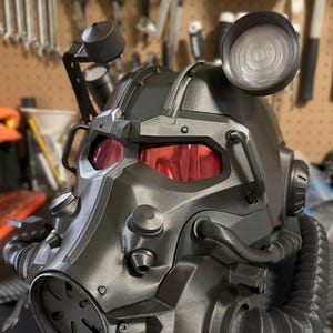 Può includere: Un casco futuristico grigio scuro con lenti colorate di rosso. Il casco presenta molteplici accessori, tra cui un riflettore, tubi e prese d'aria circolari. Il design suggerisce un tema post-apocalittico o di fantascienza.