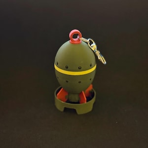 May include: A novelty keychain shaped like a cartoon bomb. The olive-green body has a yellow stripe and red accents. The keychain includes a small key and a red loop at the top.