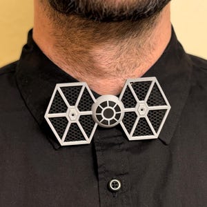 May include: A unique bow tie featuring a TIE fighter design. The bow tie has a white frame with a black mesh interior and a central circular detail. The bow tie is worn on a black collared shirt.