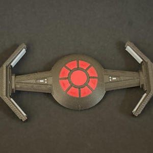 May include: A 3D-printed model of a TIE fighter, a fictional spacecraft from a science fiction franchise. The model is primarily dark gray with red accents on the central sphere. The wings extend outward from the sphere.