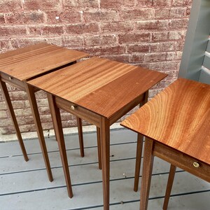 May include: A set of three matching wooden side tables, each featuring a square top with a visible wood grain pattern, four slender tapered legs, and a single small drawer equipped with a small, round, metallic knob centered on the front.