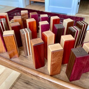 May include: A collection of rectangular wooden salt and pepper shakers featuring various natural wood grains and colors including deep red, purple, light tan, and dark brown. Each shaker has a flat top with small holes for dispensing seasoning.