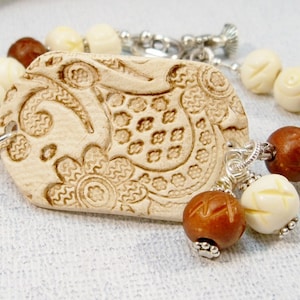 May include: A white bracelet with a carved bone charm featuring a paisley design. The bracelet also includes brown and white wooden beads and silver accents.