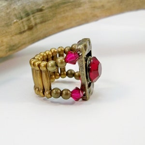 May include: A gold ring with a square-shaped setting featuring a large red gemstone. The ring is made with gold-colored beads and has a stretchy band.