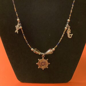 May include: A silver necklace with a blue and gold beaded chain. The necklace has three charms: a fish, a mermaid, and a ship's wheel.
