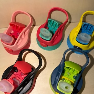 May include: Five circular water bottle lids with integrated carrying loops and flip-top spouts. The lids feature various color combinations including pink, red and teal, yellow and blue, black and red, and navy blue with lime green accents.
