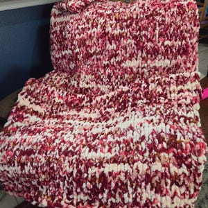 May include: A chunky knit throw blanket featuring a variegated color pattern of deep burgundy, bright pink, and cream white yarn. The blanket is constructed with thick, soft, plush loops in a consistent hand-knitted texture throughout the piece.