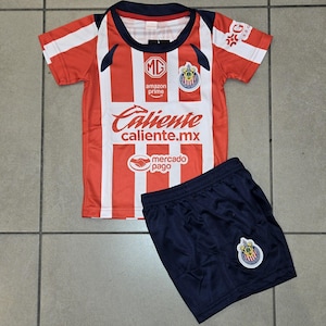 Può includere: Un completo composto da maglia da calcio a righe rosse e bianche e pantaloncini blu navy. La maglia presenta il testo "Caliente caliente.mx" e "amazon prime". I pantaloncini hanno un logo della squadra.