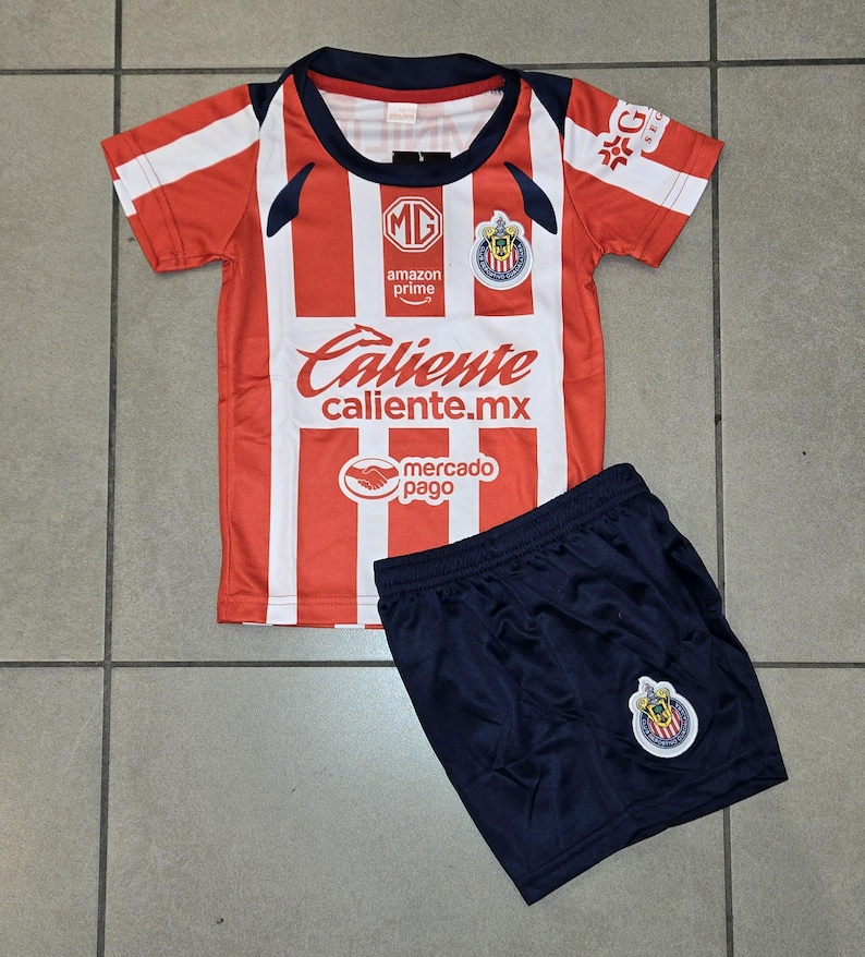 May include: A red and white striped soccer jersey and navy blue shorts set. The jersey has the text "Caliente caliente.mx" and "amazon prime" printed on it. The shorts have a team logo on the leg.