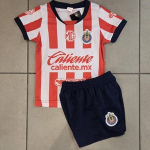 May include: A red and white striped soccer jersey with a blue collar and short sleeves. The jersey has a team logo on the left chest and the text "Caliente caliente.mx" on the front. The jersey is paired with navy blue soccer shorts with a team logo on the right leg.