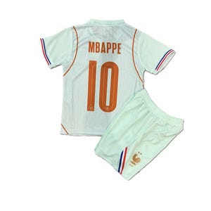 May include: A light mint green football kit consisting of a jersey and shorts. The jersey displays the name MBAPPE and the number 10 in orange on the back, with orange piping and blue, white, and red sleeve trim. The shorts feature a gold rooster emblem.