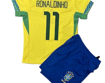 Puede incluir: Una camiseta de fútbol amarilla con detalles en verde, que presenta el nombre RONALDINHO y el número 11 en verde oscuro en la espalda, junto con unos pantalones cortos de fútbol azules que muestran el escudo CBF Brasil con cinco estrellas.