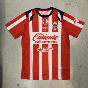 May include: Red and white striped soccer jersey with the text "Caliente" and "caliente.mx". The jersey also features the logos of "MG", "Amazon Prime", and "mercado pago". The sleeves are red with white accents.