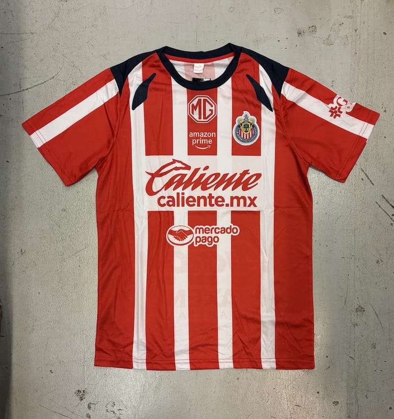 May include: Red and white striped soccer jersey with the text "Caliente" and "caliente.mx". The jersey also features the logos of "MG", "Amazon Prime", and "mercado pago". The sleeves are red with white accents.