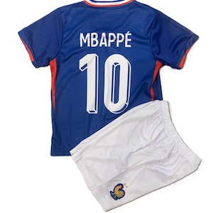 Può includere: Una maglia da calcio blu con inserti rossi sui lati e colletto bianco, che riporta il nome MBAPPÉ e il numero 10 in bianco sul retro, abbinata a pantaloncini sportivi bianchi con un piccolo emblema di un gallo blu e oro sulla gamba.
