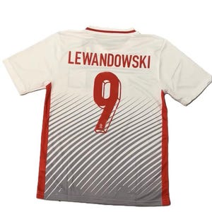 May include: A white short-sleeved sports jersey featuring the name LEWANDOWSKI and the number 9 in red on the back. The lower portion of the jersey displays a gray diagonal striped pattern, with solid red vertical panels along both side seams.