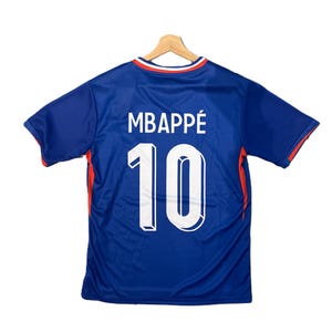 May include: A blue short-sleeved sports jersey featuring the name MBAPPÉ and the number 10 printed in white on the back. The collar has red and white trim, and there are red accents along the sides of the shirt, which is displayed on a hanger.