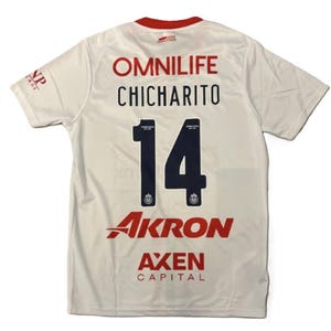 Peut inclure: Un maillot de sport blanc à manches courtes avec un col rouge. Le dos affiche le texte OMNILIFE, CHICHARITO, le numéro 14 en bleu foncé, ainsi que les logos rouges AKRON et AXEN CAPITAL. Un petit logo NP est visible sur la manche droite.