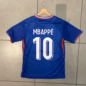 K&ouml;nnte beinhalten: Ein blaues Fu&szlig;balltrikot mit dem Namen "MBAPP&Eacute;" und der Nummer "10" in Wei&szlig;. Das Trikot hat rote und wei&szlig;e Details an den &Auml;rmeln und am Kragen. Es h&auml;ngt an einem Holzb&uuml;gel.