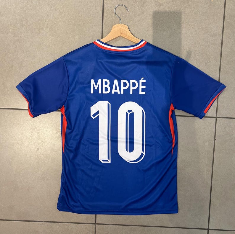 K&ouml;nnte beinhalten: Ein blaues Fu&szlig;balltrikot mit dem Namen "MBAPP&Eacute;" und der Nummer "10" in Wei&szlig;. Das Trikot hat rote und wei&szlig;e Details an den &Auml;rmeln und am Kragen. Es h&auml;ngt an einem Holzb&uuml;gel.