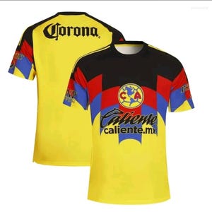 Peut inclure: Maillot de football jaune avec des détails noirs et rouges. Le maillot porte le mot "Corona" au dos et le logo "Club America" sur le devant. Le texte "Caliente caliente.mx" est également visible.