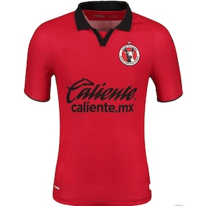 Pode incluir: Camisola de futebol vermelha com guarnições pretas e um logotipo de equipa preto com um cão na frente. O texto "Caliente caliente.mx" está impresso na frente da camisola.