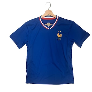 K&ouml;nnte beinhalten: Ein k&ouml;nigsblaues Kurzarmtrikot mit V-Ausschnitt und rot-wei&szlig;em Besatz. Auf der Brust befindet sich ein gesticktes goldenes Hahn-Emblem &uuml;ber den Buchstaben FFF und zwei kleinen Sternen. Das Trikot wird auf einem Holzkleiderb&uuml;gel pr&auml;sentiert.