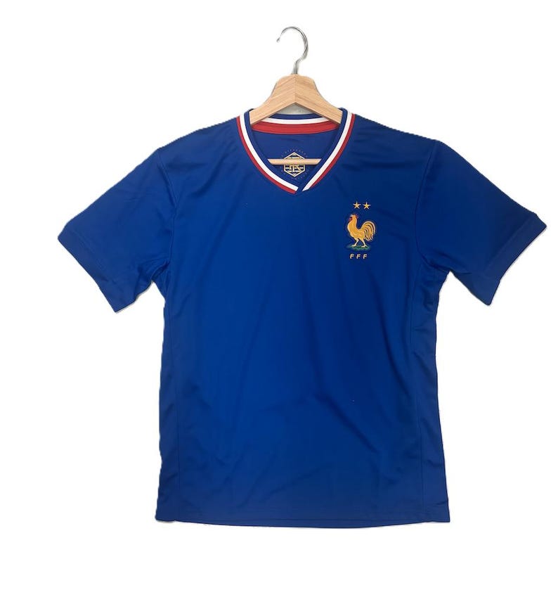 K&ouml;nnte beinhalten: Ein k&ouml;nigsblaues Kurzarmtrikot mit V-Ausschnitt und rot-wei&szlig;em Besatz. Auf der Brust befindet sich ein gesticktes goldenes Hahn-Emblem &uuml;ber den Buchstaben FFF und zwei kleinen Sternen. Das Trikot wird auf einem Holzkleiderb&uuml;gel pr&auml;sentiert.