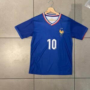 K&ouml;nnte beinhalten: Ein blaues Fu&szlig;balltrikot mit der Nummer 10 in Wei&szlig; auf der Vorderseite. Das Trikot hat einen V-Ausschnitt mit rotem und wei&szlig;em Besatz und ein Hahn-Emblem auf der Brust. Das Trikot h&auml;ngt an einem Holzb&uuml;gel.