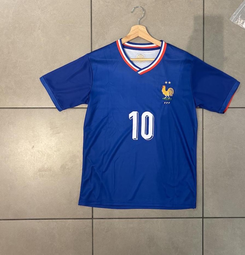 K&ouml;nnte beinhalten: Ein blaues Fu&szlig;balltrikot mit der Nummer 10 in Wei&szlig; auf der Vorderseite. Das Trikot hat einen V-Ausschnitt mit rotem und wei&szlig;em Besatz und ein Hahn-Emblem auf der Brust. Das Trikot h&auml;ngt an einem Holzb&uuml;gel.