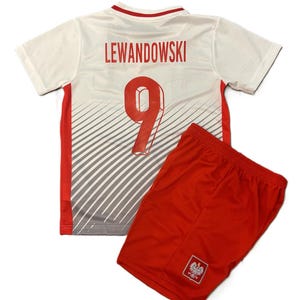 May include: A white short-sleeved jersey with red accents, featuring the name LEWANDOWSKI and the number 9 in red on the back, paired with red athletic shorts that display a small emblem of a white eagle on a red shield on the lower leg.