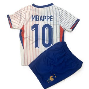 May include: A white football shirt with thin red vertical pinstripes, blue trim, and the name MBAPPÉ with the number 10 in blue. Paired with blue athletic shorts featuring a small yellow cockerel emblem and two gold stars above the letters FFF.