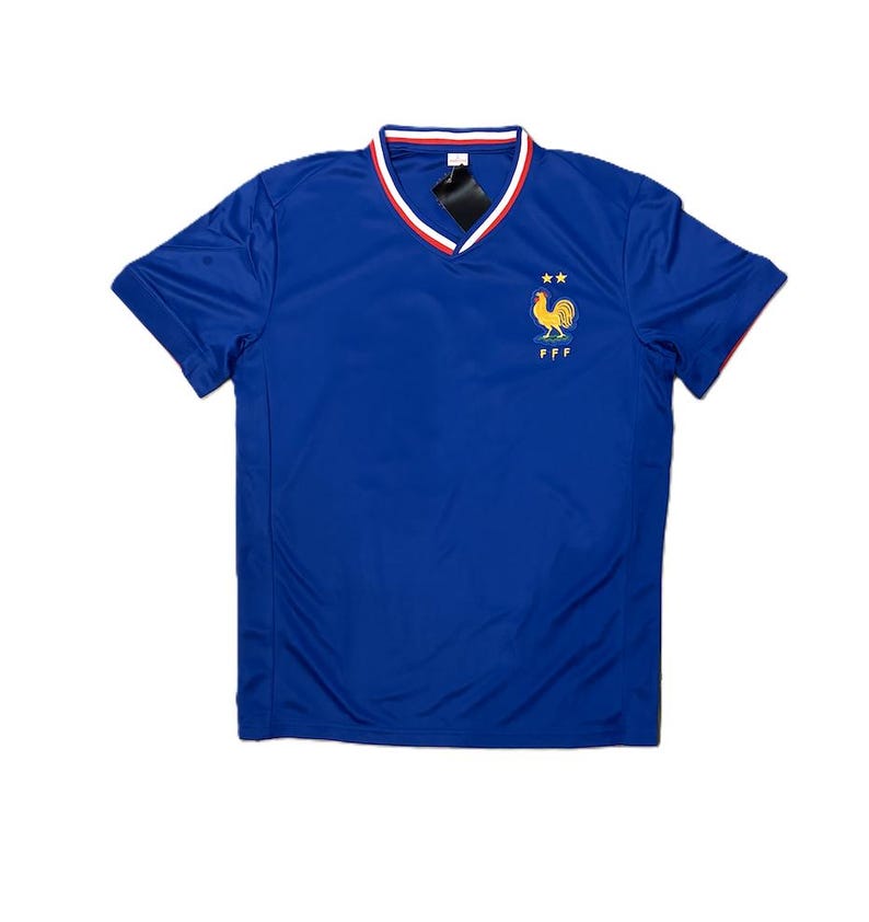May include: A royal blue short-sleeved jersey with a V-neck collar featuring red and white trim. The chest displays an embroidered yellow rooster emblem above the letters FFF and two small yellow stars positioned above the rooster icon.
