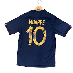 May include: A dark blue short-sleeved jersey featuring the name MBAPPE in yellow lettering above the number 10. The number 10 is filled with small photographic images of a player. The sleeve cuffs have a red, white, and blue striped detail.