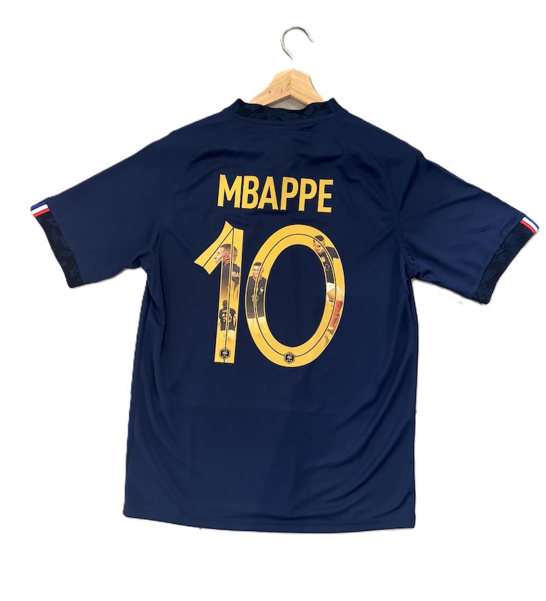 May include: A dark blue short-sleeved jersey featuring the name MBAPPE in yellow lettering above the number 10. The number 10 is filled with small photographic images of a player. The sleeve cuffs have a red, white, and blue striped detail.