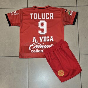 May include: A red soccer jersey and shorts set. The jersey has the word "TOLUCA" and the number "9" in white. The name "A. VEGA" and the word "Calient" are also visible. The shorts are red with a gold emblem.