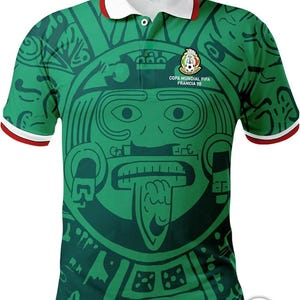 Może przedstawiać: Zielona koszulka polo z białym kołnierzykiem i czerwonym wykończeniem. Koszulka ma duży, inspirowany azteckimi wzorami wzór z twarzą w środku. Na koszulce nadrukowany jest tekst "COPA MUNDIAL FIFA FRANCIA 98".