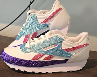 May include: White Reebok sneakers with blue, pink, and purple rhinestone embellishments. The shoes feature a white sole and laces, with the Reebok logo visible on the side. The design includes a purple toe cap and a blue and pink stripe.
