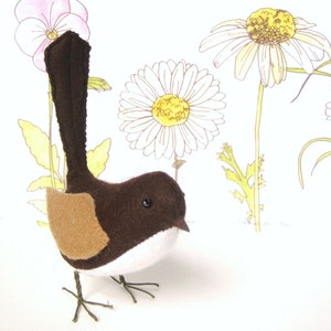 Könnte beinhalten: Ein brauner und weißer Filzvogel mit langem Schwanz, der auf einem Drahtständer sitzt. Der Vogel ist von Aquarell-Illustrationen von Blumen umgeben.