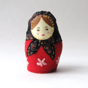 May include: A red felt nesting doll with a black and white patterned headscarf. The doll has a hand-stitched face with brown hair and pink cheeks.