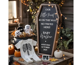 May include: A wooden coffin-shaped sign with white text reading OUR little treat IS ON THE WAY NO TRICKS HERE! BABY WALKER ARRIVING OCTOBER 2025. A white baby onesie with a black skeleton ribcage print, a black cauldron, and small pumpkins.