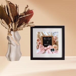 May include: A black picture frame with a white mat and a photo of a wooden sign that says "It's a Girl" with a pink bow and a sonogram photo. The photo is surrounded by pink flowers and a white fluffy background.